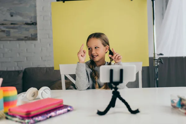 Smiling girl pointing with fingers while recording video blog on smartphone at home on yellow background — Stock Photo