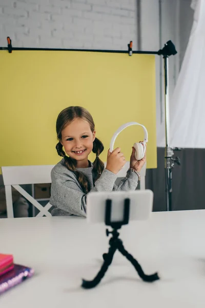 Sorrindo blogueiro pré-adolescente mostrando fones de ouvido sem fio perto de telefone celular borrado no fundo amarelo em casa — Fotografia de Stock