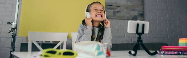 Excited video blogger in wireless headphones near blurred smartphone on holder, banner — Stock Photo