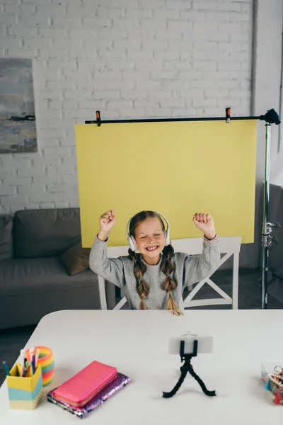 Excited video blogger in wireless headphones gesturing near cellphone and yellow background at home — Stock Photo