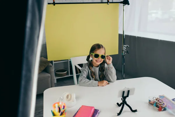 Sonriente video blogger demostrando gafas de sol de moda cerca de varios accesorios y teléfono móvil borroso - foto de stock