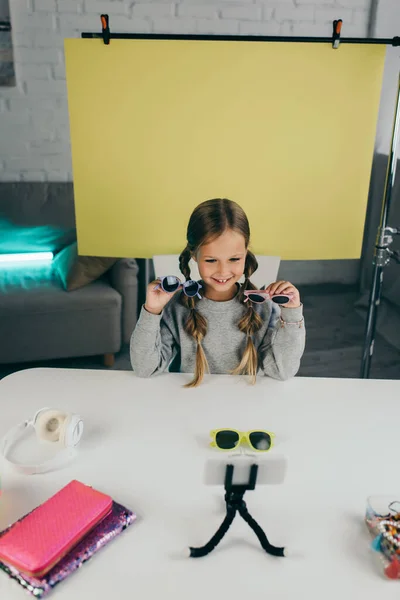 Blogger sonriente mostrando gafas de sol elegantes frente a un teléfono inteligente borroso cerca del fondo amarillo en casa - foto de stock