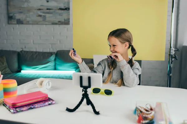 Щасливий блогер, що показує стильні сонцезахисні окуляри біля розмитих мобільних телефонів та пенали з навушниками на столі — стокове фото
