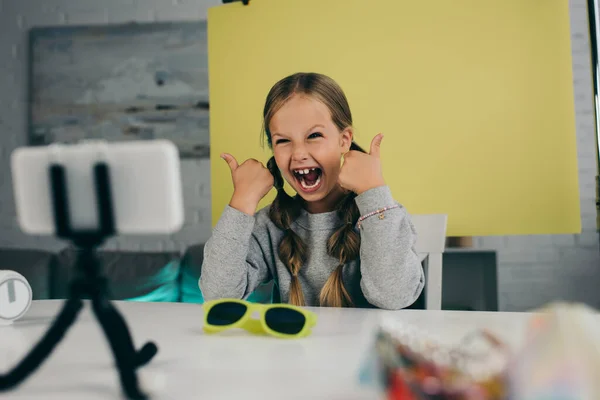 Blogueur vidéo excité montrant pouces vers le haut près de lunettes de soleil à la mode et le téléphone mobile flou sur le titulaire — Photo de stock