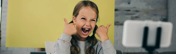 Fille étonnée avec bouche ouverte montrant pouces vers le haut près de téléphone cellulaire flou sur fond jaune à la maison, bannière — Photo de stock