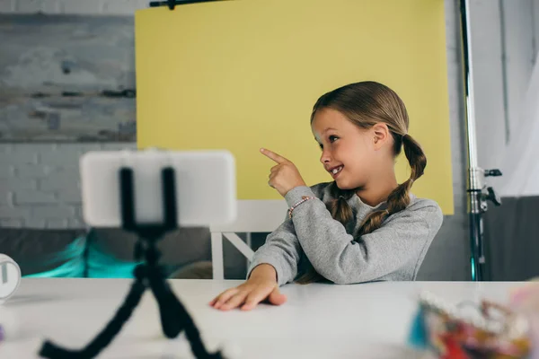 Smiling girl pointing with finger on yellow background near blurred cellphone on phone holder — Stock Photo