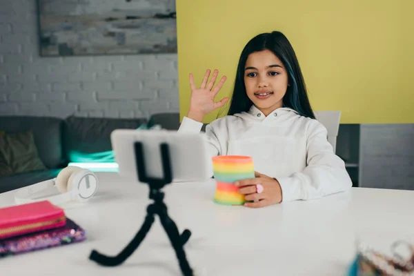 Preteen video blogger with spiral toy waving hand near blurred cellphone on phone holder — Stock Photo