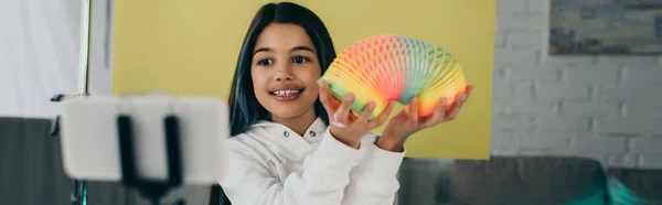 Cheerful video blogger showing spiral toy near blurred mobile phone at home, banner — Stock Photo