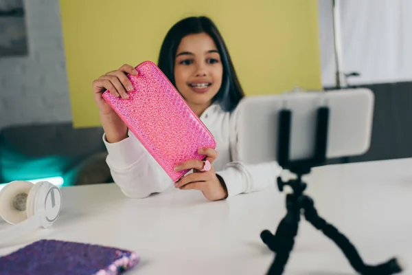 Smiling blurred girl showing shiny pencil case near smartphone on phone holder — Stock Photo