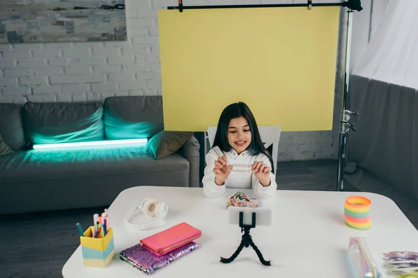Sonriente chica sosteniendo pulsera de cuentas durante la grabación de vídeo blog en el teléfono inteligente en casa - foto de stock