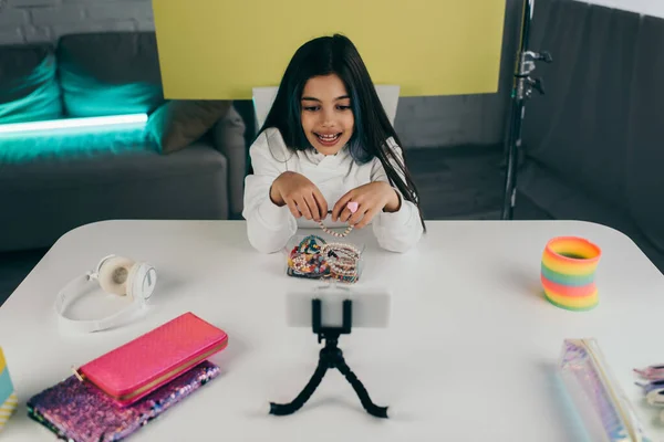 Fröhlicher Videoblogger spricht in der Nähe von Perlenarmbändern und Handy auf Halterung — Stockfoto