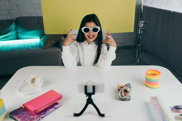 Menina morena feliz demonstrando óculos de sol elegantes perto de smartphone borrado e fundo amarelo em casa — Fotografia de Stock