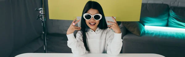 Усміхнений блогер демонструє модні сонцезахисні окуляри на жовтому тлі вдома, банер — стокове фото