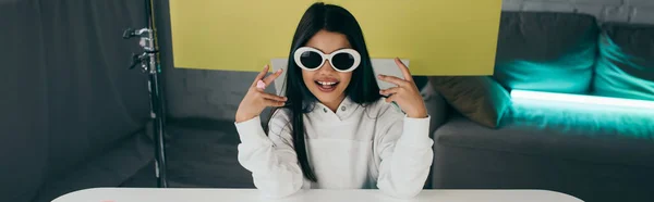 Blogueur souriant démontrant des lunettes de soleil à la mode sur fond jaune à la maison, bannière — Photo de stock