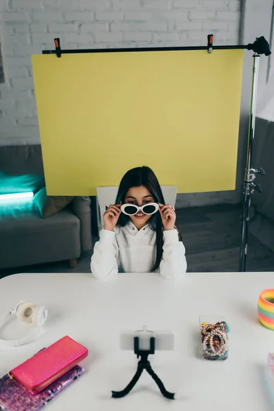 Video blogger showing trendy sunglasses near blurred smartphone and yellow background with copy space at home — Stock Photo