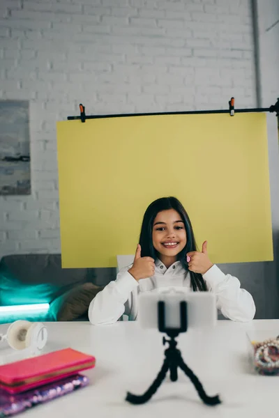 Щасливий відео блогер показує великі пальці на жовтому тлі біля мобільного телефону вдома — стокове фото
