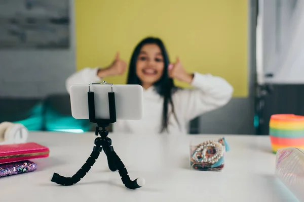 Selective focus of smartphone on holder near beaded bracelets and blurred video blogger showing thumbs up — Stock Photo
