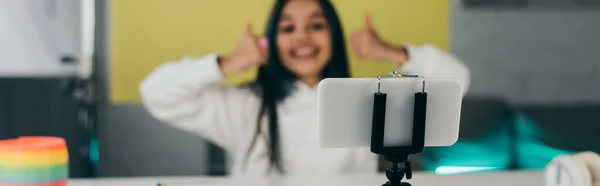 Alegre video blogger mostrando pulgares hacia arriba cerca de teléfono inteligente en el titular del teléfono sobre fondo borroso, bandera - foto de stock