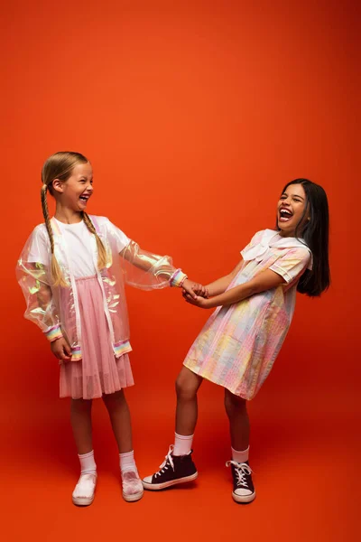 Excited brunette girl holding hand of friend and laughing on orange background — Stock Photo