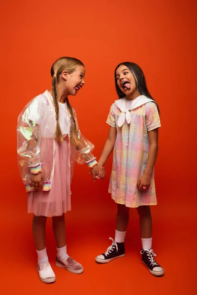 Full length of funny girls holding hands and sticking out tongues at each other on orange background — Stock Photo