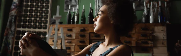 Profile of thoughtful african american woman sitting in workshop and looking away, banner — Stock Photo