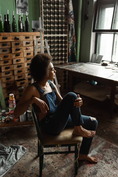 Красивая и мечтательная американская художница в комбинезоне сидит босиком в мастерской и смотрит в окно — стоковое фото