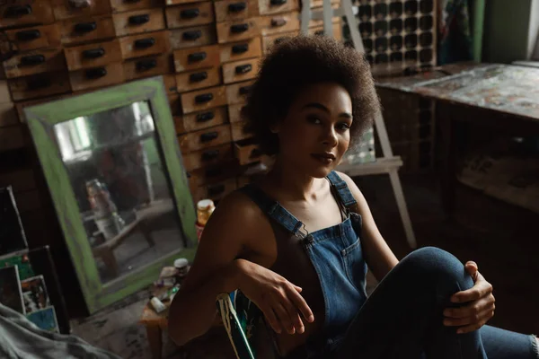 African american woman in overalls sitting in art workshop near vintage mirror and wooden furniture — Stock Photo