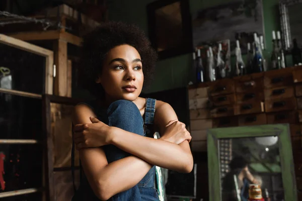 Pensosa donna afroamericana seduta con le braccia incrociate in studio d'arte vicino specchio d'epoca su sfondo sfocato — Foto stock