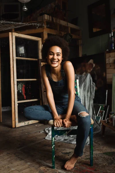 Mulher americana africana descalça em macacão sentado no estúdio de arte e olhando para a câmera — Fotografia de Stock