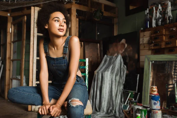 Joven mujer afroamericana en overol denim mirando hacia otro lado en taller de arte cerca de latas con pintura - foto de stock