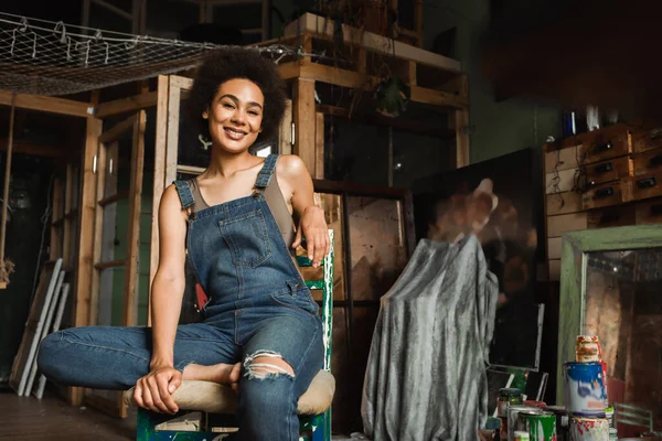 Africano americano donna in denim tuta seduta in arte studio e sorridente a macchina fotografica — Foto stock