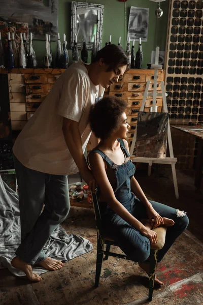 Молодая босиком художница, стоящая рядом с сексуальной африканской женщиной в джинсовом комбинезоне, сидит в мастерской — стоковое фото