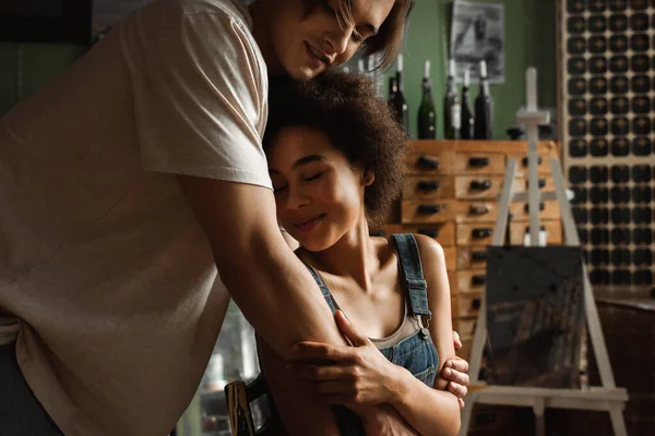 Artiste souriant étreignant jolie femme afro-américaine assise dans un atelier les yeux fermés — Photo de stock