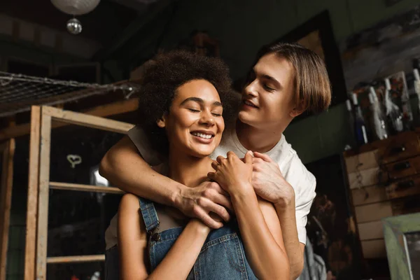 Feliz artista abrazando bastante afroamericana novia sonriendo con los ojos cerrados en el taller - foto de stock