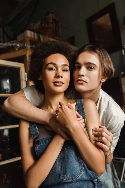 Young interracial couple embracing and looking at camera in art studio — Stock Photo