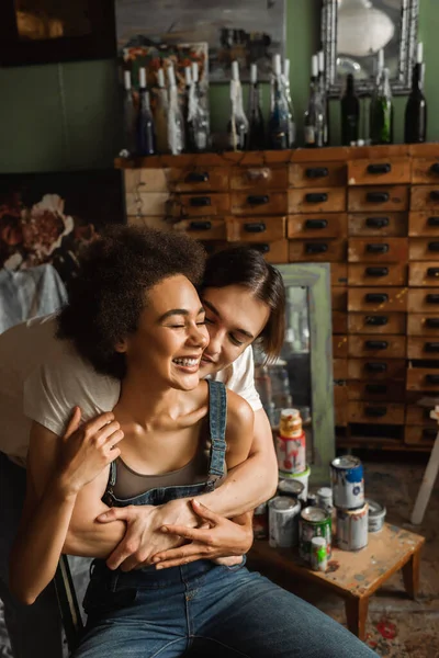 Giovane artista che abbraccia allegra donna afroamericana vicino mobili vintage in laboratorio — Foto stock