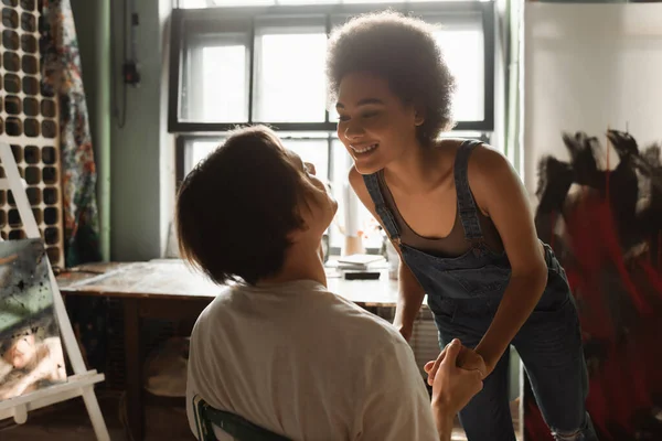 Happy multiethnic artists holding hands and looking at each other in workshop — Stock Photo