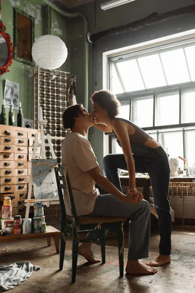 Side view of barefoot african american woman in overalls near boyfriend sitting in workshop with large window — Stock Photo