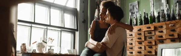 Молодая межрасовая пара художников обнимаются, сидя в мастерской и глядя в большое окно, баннер — стоковое фото