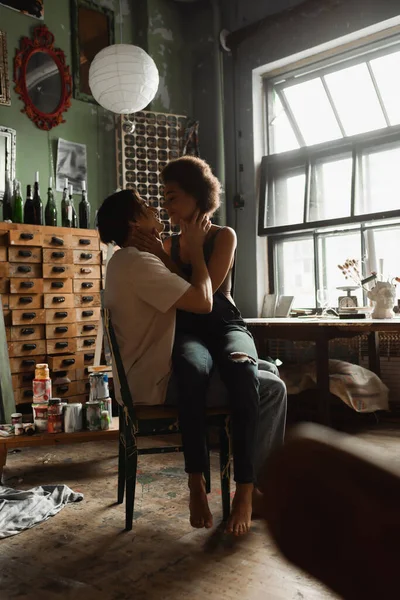 Sexy african american woman in overalls sitting on laps of boyfriend in art studio with vintage furniture and large window — Stock Photo