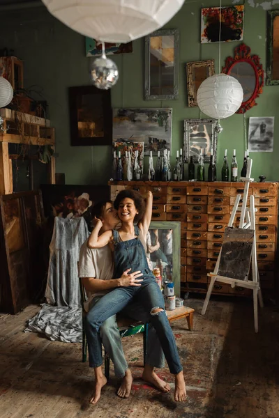 Веселая межрасовая пара, обнимающаяся на стуле в художественной студии со старинной мебелью и зеркалами — стоковое фото