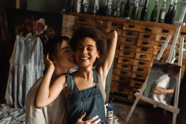 Cheerful african american woman with closed eyes near man hugging her in art studio — Stock Photo