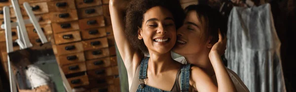 Joven artista cerca de mujer afroamericana sonriendo con los ojos cerrados en el taller, pancarta - foto de stock