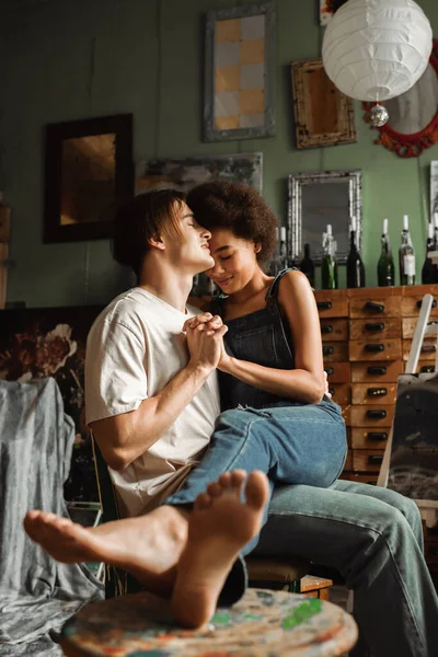 Barfüßige Afroamerikanerin in Jeans-Overalls hält Händchen mit Mann, während er auf seinem Schoß in der Werkstatt sitzt — Stockfoto