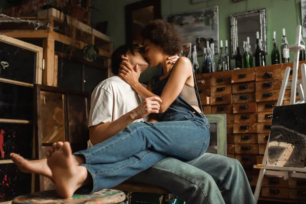 Descalzo afroamericana mujer en denim overoles besándose con joven artista en taller - foto de stock