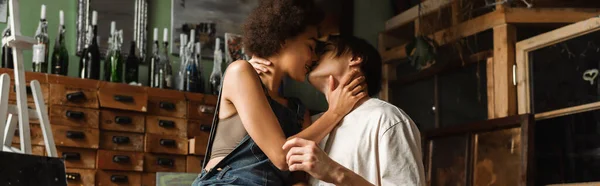 Side view of young interracial couple kissing in workshop near vintage furniture and bottles with candles, banner — Stock Photo