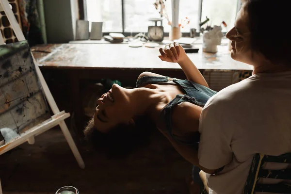 Young artist holding cheerful and sexy african american girlfriend while sitting on chair in workshop — Stock Photo