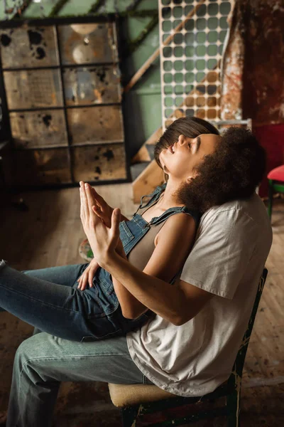 Seitenansicht einer lächelnden und sinnlichen afrikanisch-amerikanischen Frau, die in der Werkstatt auf dem Schoß ihres Freundes sitzt — Stockfoto