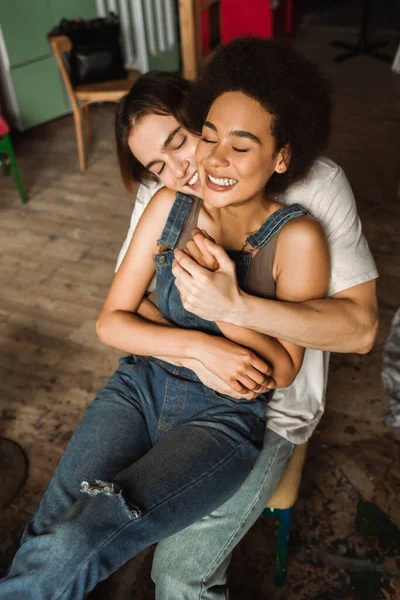 High angle view of young man hugging smiling african american girlfriend in overalls in art studio — Stock Photo