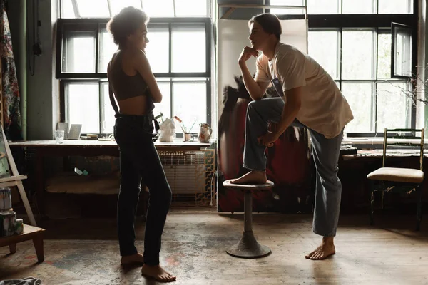 Full length of young barefoot artist looking at sexy african american woman in workshop with large windows — Stock Photo